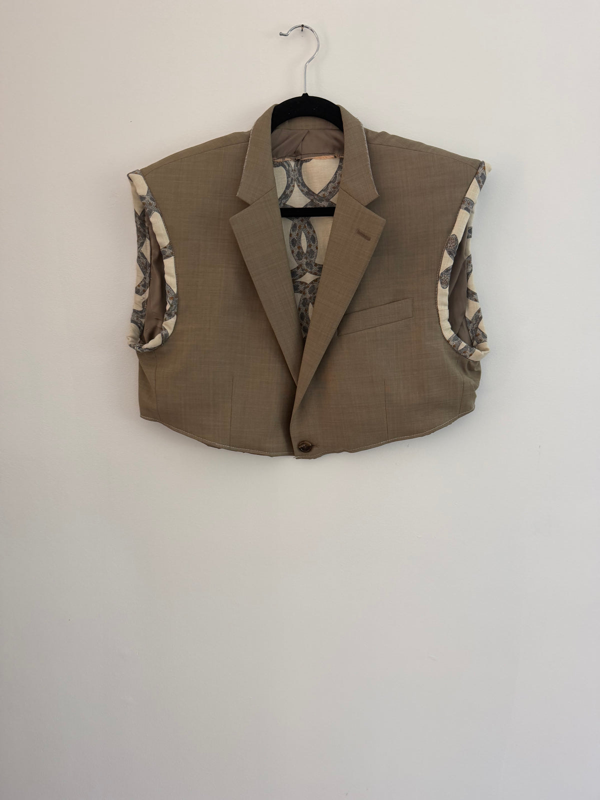 A cropped taupe-brown blazer vest with patterned lining, hanging on a black hanger against a plain white wall.
